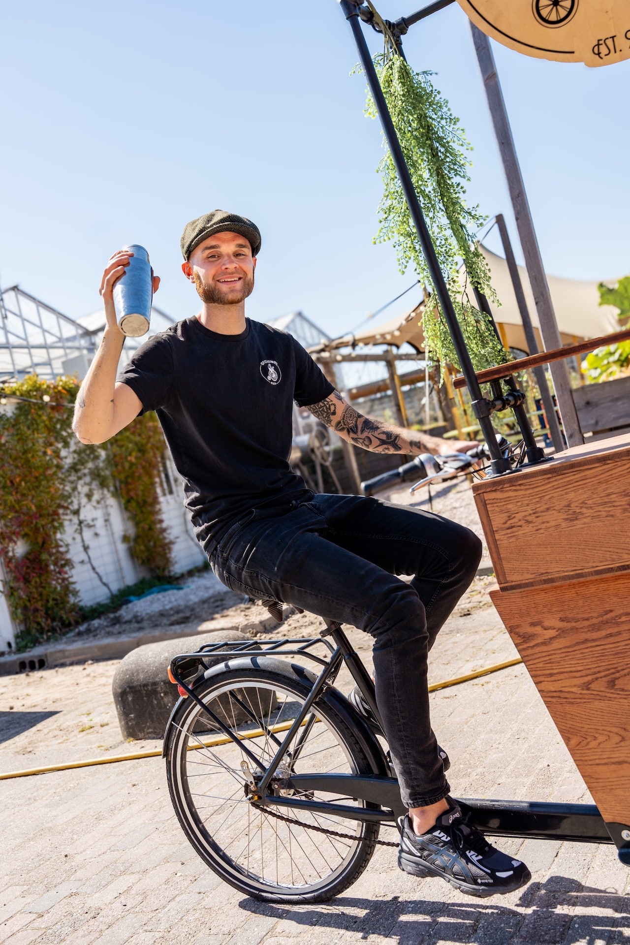 Bruid en bruidegom proosten met rode cocktails bij De Cocktailfiets-bakfietsbar in parksetting, met bartender die glimlachend toekijkt